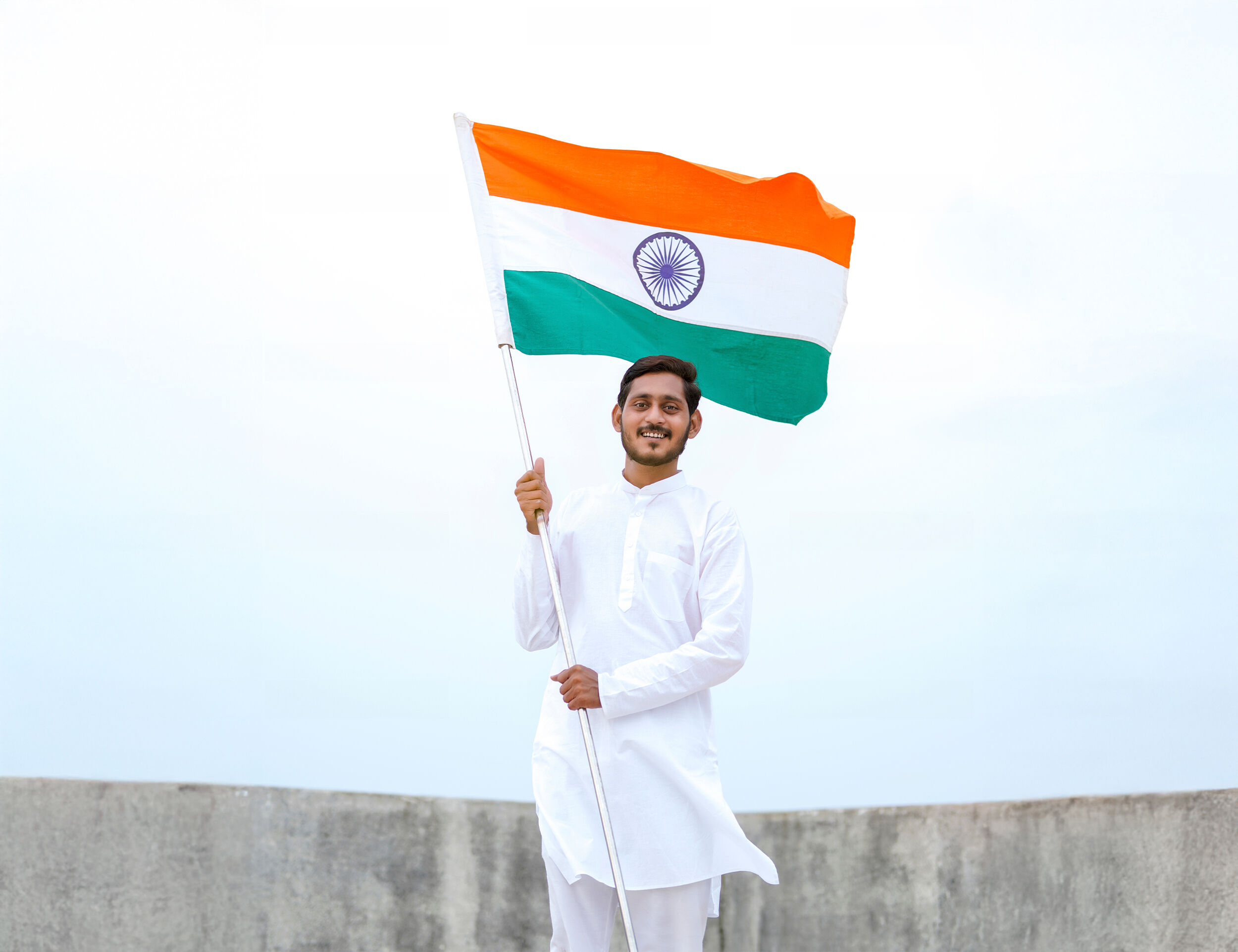 Young man in traditional white kurta proudly holding the Indian national flag against a clear sky, celebrating Independence Day.